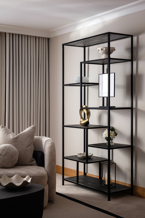 Black metal-framed open shelving unit styled with sculptural objects, a framed print and a brass figure, beside a cream boucle armchair and round black coffee table. Designed by Lisa Hensby Design & Build Studio.