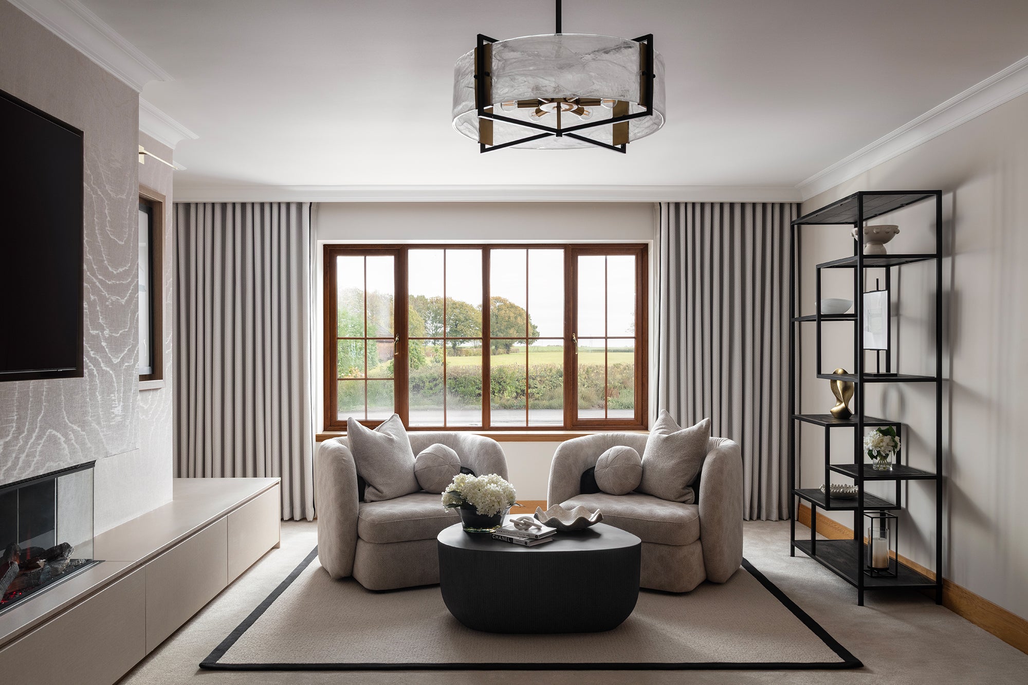 Reading corner with two sculptural cream armchairs framing a round black coffee table, set against a large window, with a media wall and black metal shelving unit to either side.