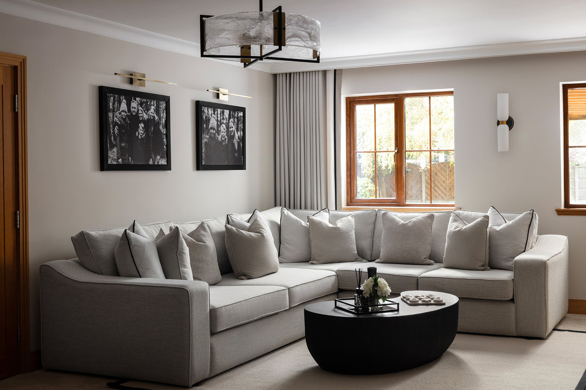 Large grey L-shaped corner sofa layered with neutral cushions, a round black coffee table and monochrome framed prints, in a soft neutral living room with a statement glass pendant. Designed by Lisa Hensby of Lisa Hensby Design & Build Studio.